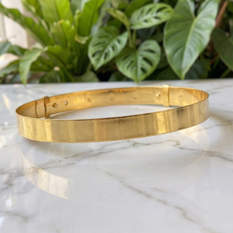 A polished gold metal bangle bracelet with a flat, wide band and a spring-ring clasp, resting on a white marble surface with green plants in the background.