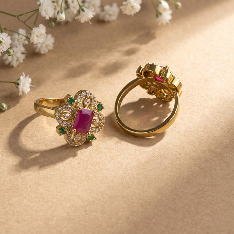 Two gold rings displayed on a beige surface with soft shadows and small white flowers in the background. One ring shows a rectangular pink gemstone surrounded by clear stones and small green accents, and the other shows the back of the ring with a matching pink stone.
