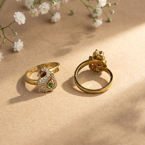 Two gold rings displayed on a beige surface with soft shadows and small white flowers in the background. One ring is shaped like a peacock with a clear crystal body, a green emerald, and a red ruby accent.