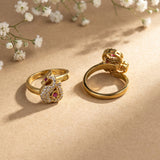 Two gold rings displayed on a beige surface with small white flowers in the background. One ring features a swan-shaped setting paved with clear stones and accented with red gemstones, and the other shows the back of the ring with a crown-like detail and red gemstone.