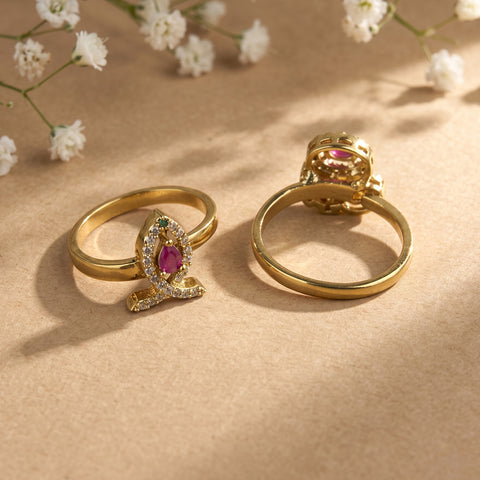 Two gold rings displayed on a beige surface with small white flowers in the background. One ring shows a leaf-shaped setting with a pink gemstone and small clear stones, and the other shows the back of the ring with a matching pink stone and decorative top.