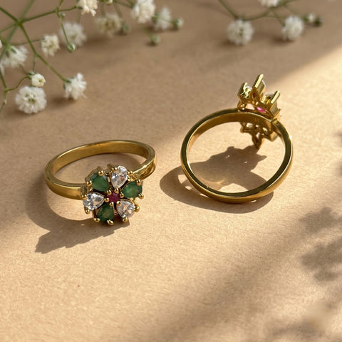 Two gold rings displayed on a beige surface with soft shadows and small white flowers in the background. One ring shows a floral cluster with green stones, clear stones, and a small red center stone.