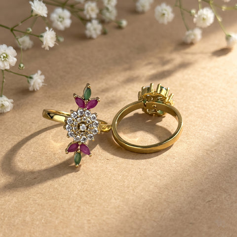 A pair of gold rings displayed on a textured beige surface with soft white baby's breath flowers in the background. One ring is shown from the front, featuring a floral cluster with clear stones and pink gemstones, while the other is shown from the back, displaying its setting.