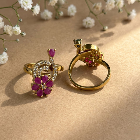 Two gold rings with pink gemstone floral and musical note details, displayed on a textured beige surface with small white flowers in the background.