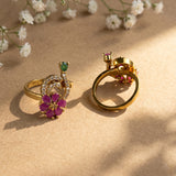 Two gold rings displayed on a beige surface with soft shadows and small white flowers in the background. One ring features a purple flower with pink centers, a green stone, and white crystal crescent details, while the other shows the back with a pink gemstone and setting.