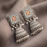 A pair of ornate silver jhumka earrings with square floral enamel panels and dangling pearl beads, displayed on a draped taupe fabric background.