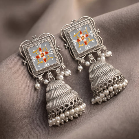 A pair of ornate silver jhumka earrings with square floral enamel panels and dangling pearl beads, displayed on a draped taupe fabric background.
