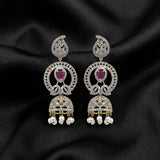 A pair of ornate Indian jhumka earrings with silver-toned metal, clear pavé stones, oval ruby gemstones, and dangling pearl-like beads, displayed on a black satin fabric background.
