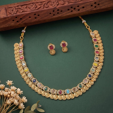 Gold-toned traditional necklace and matching stud earrings with multiple colorful gemstone accents, arranged on a dark green surface with a carved wooden prop and small dried flowers.
