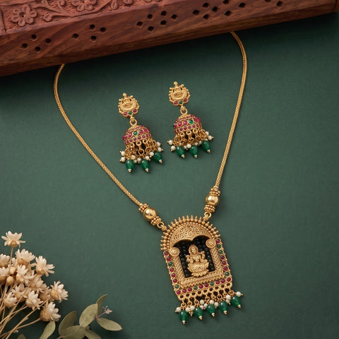 Gold-toned traditional Indian jewelry set featuring a necklace with a detailed arch-shaped pendant depicting a seated deity, accompanied by matching drop earrings. The jewelry is embellished with red stones, green bead drops, and small white pearl-like accents, arranged on a dark green background with decorative dried flowers and a carved wooden prop.