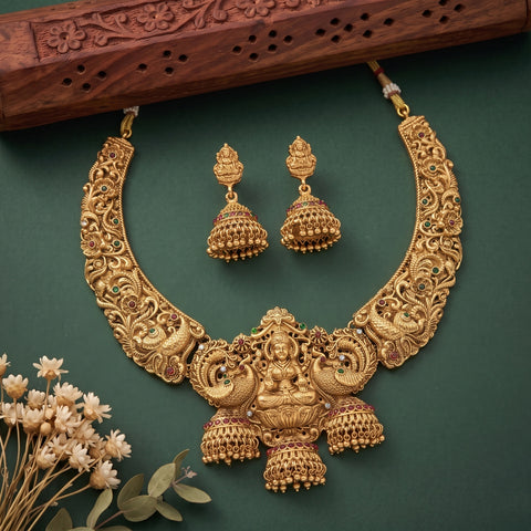 An ornate gold-toned traditional necklace and matching jhumka earrings displayed on a dark green background, with a carved wooden prop at the top and small dried flowers at the bottom left.