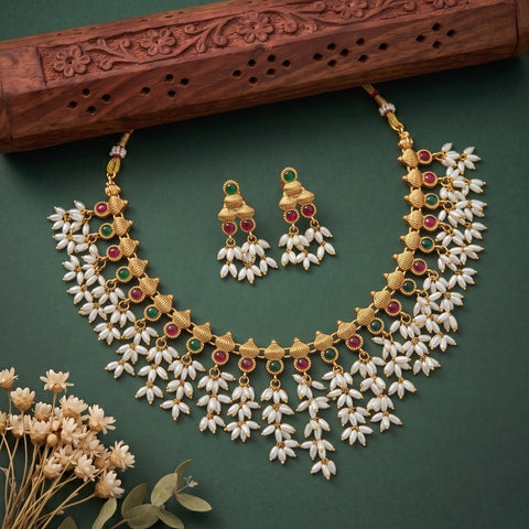Gold-plated traditional Indian jewelry set featuring a necklace and matching earrings, decorated with white pearl-like flowers and accented with red and green stones, displayed on a dark green background with a carved wooden prop.