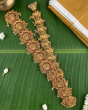 A pair of ornate gold-toned traditional Indian jewelry pieces, likely a necklace and matching bracelet or earrings, decorated with intricate filigree, red stone accents, and small pearl-like beads, arranged diagonally on a large green banana leaf with white flowers and a folded yellow textile in the background.