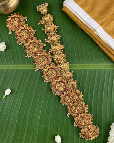 A pair of ornate gold-toned traditional Indian jewelry pieces, likely a necklace and matching bracelet or earrings, decorated with intricate filigree, red stone accents, and small pearl-like beads, arranged diagonally on a large green banana leaf with white flowers and a folded yellow textile in the background.