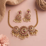 Gold-toned Indian bridal jewelry set featuring a choker necklace, matching earrings, and a chain, embellished with gold-plated elements, white faux pearls, and deep pink stones, displayed on a dusty pink background with decorative burlap and a pink flower.
