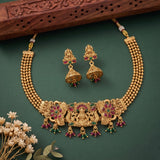 Gold-toned traditional Indian jewelry set featuring a choker necklace with a central deity motif and floral inlays, matching jhumka earrings, and decorative dried flowers on a dark green background.