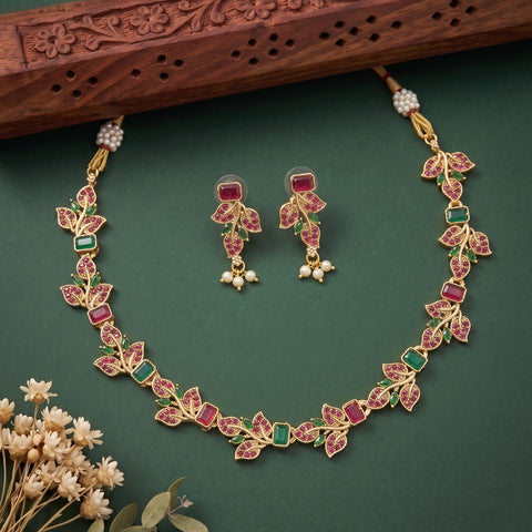 Gold-plated Indian jewelry set featuring a necklace and matching earrings with red and green stone inlay, displayed on a dark green background with a carved wooden prop and small dried flowers.