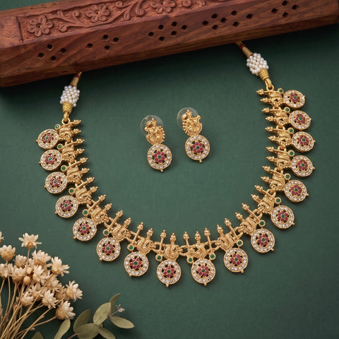 Gold-toned traditional necklace and matching stud earrings with circular motifs, red stone centers, and small green stone accents, arranged on a dark green background with decorative dried flowers and a carved wooden prop.