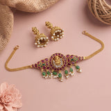 Gold-toned traditional Indian jewelry set featuring a choker necklace, matching earrings, and a chain, decorated with green stones, white pearls, and a central gold motif, displayed on a pink background with burlap and a flower prop.