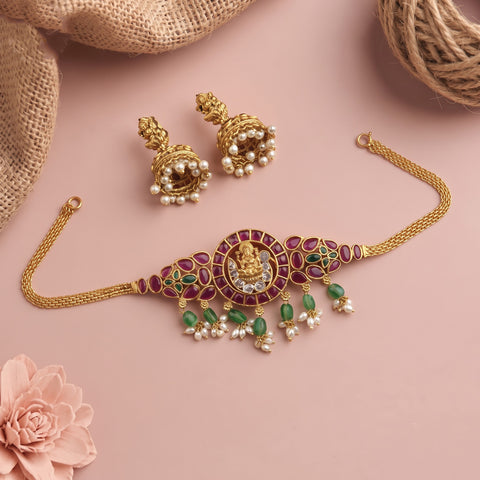 Gold-toned traditional Indian jewelry set featuring a choker necklace, matching earrings, and a chain, decorated with green stones, white pearls, and a central gold motif, displayed on a pink background with burlap and a flower prop.