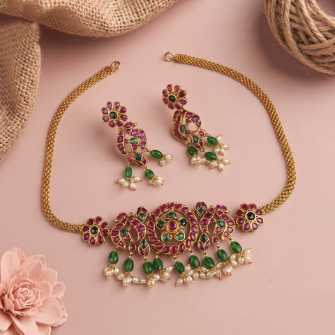 Gold-plated jewelry set featuring a necklace and matching earrings adorned with pink stones, green stones, and white faux pearls, styled on a pink background with burlap and a flower prop.