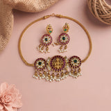 Gold-toned traditional Indian jewelry set featuring a necklace and matching earrings with red stone accents, green stones, and white pearl-like drops, arranged on a pink background with burlap and a flower prop.