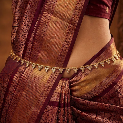 A close-up of a person wearing a traditional Indian saree in deep maroon and gold, accessorized with a gold chain waist belt decorated with small clear stones.