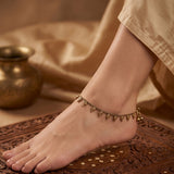A close-up of a person's foot and ankle wearing a delicate gold-toned anklet with small dangling bead-like charms, styled with beige fabric and a carved wooden surface.