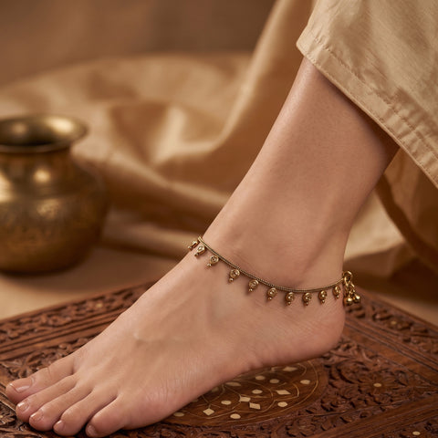 A close-up of a person's foot and ankle wearing a delicate gold-toned anklet with small dangling bead-like charms, styled with beige fabric and a carved wooden surface.