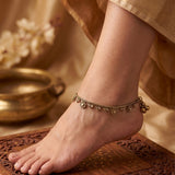 A close-up of a person's foot and ankle wearing an ornate gold-toned anklet with dangling floral and bead details, standing on a carved wooden surface with a soft beige fabric and brass bowl in the background.