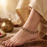 A close-up of a person's foot and ankle wearing an ornate gold-toned anklet with dangling bell charms, styled with beige fabric and a carved wooden surface.