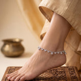 A delicate silver-toned anklet with small floral clusters and dangling beads worn on a person's ankle, shown against a warm neutral background with draped fabric and a carved wooden surface.