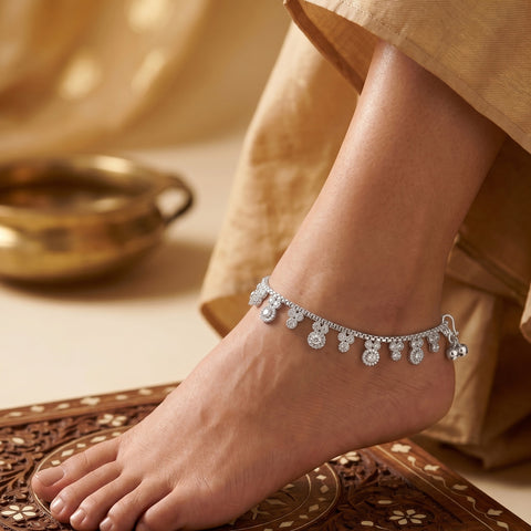 A close-up of a person's foot and ankle wearing a delicate silver-toned anklet adorned with small dangling teardrop-shaped clear stones, styled with a beige garment and a carved wooden surface.