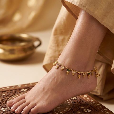 A gold-toned traditional Indian ankle chain with small hanging motifs and a few green stone accents, shown worn on a person's ankle with a beige garment and decorative mat in the background.