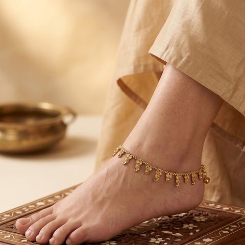A close-up of a person's bare foot wearing a delicate gold anklet with dangling decorative charms, standing on an ornate wooden surface with draped beige fabric in the background.