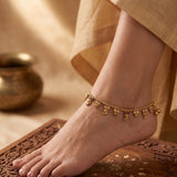 A delicate gold anklet with small dangling charms is shown worn on a person's ankle, with bare feet resting on a carved wooden surface and draped beige fabric in the background.