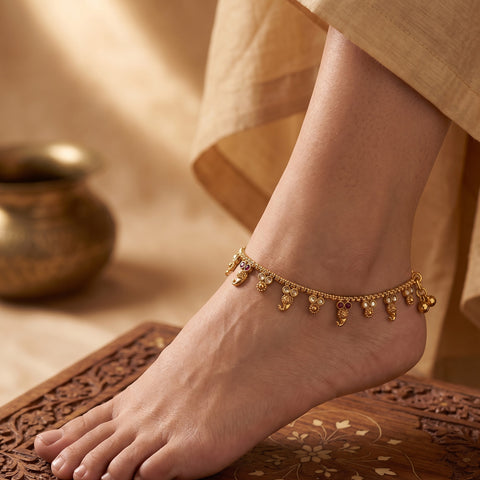 A delicate gold anklet with small dangling charms is shown worn on a person's ankle, with bare feet resting on a carved wooden surface and draped beige fabric in the background.