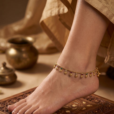 A close-up of a person's foot and ankle wearing a delicate gold chain anklet adorned with small hanging gold beads and tiny green and red stone accents, set against a warm, traditional background with brass vessels.