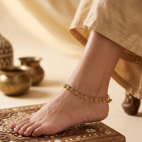 A close-up of a person's foot and ankle wearing a delicate gold anklet with small dangling motifs, standing on a carved wooden platform with traditional brass pots in the background.