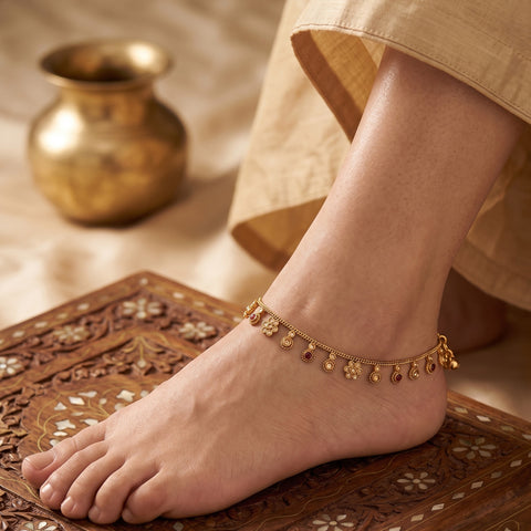 A delicate gold-toned anklet with small dangling floral and bead motifs worn on a person's ankle, shown with a foot resting on an ornate carved wooden surface. A soft beige garment is visible at the top, and a blurred brass pot appears in the background.