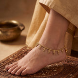 A close-up of a person's foot and ankle wearing a delicate gold chain anklet decorated with small dangling coins, standing on a carved brown mat with a beige garment draped nearby.