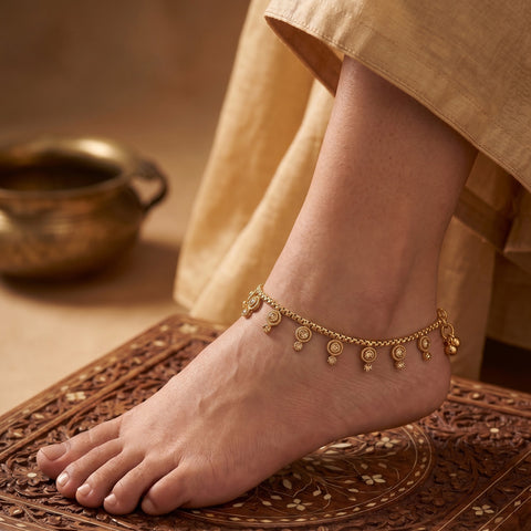 A close-up of a person's foot and ankle wearing a delicate gold chain anklet decorated with small dangling coins, standing on a carved brown mat with a beige garment draped nearby.