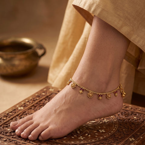 A close-up of a person's bare foot and ankle wearing a delicate gold chain anklet decorated with small hanging floral charms, standing on a carved wooden mat with a soft beige garment draped nearby.