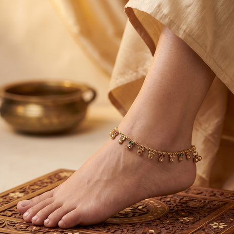 A delicate gold ankle chain bracelet with small hanging charms, worn on a person's ankle as their foot rests on an ornate carved wooden surface. The background is softly blurred with a brass bowl and warm beige fabric.