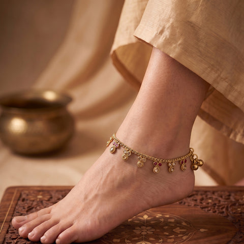 A close-up of a person's foot and ankle wearing a delicate gold chain anklet decorated with small dangling charms, set against a warm neutral background with a brass pot and patterned wooden mat.