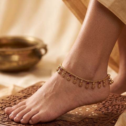 A delicate gold anklet with small coin charms and red gemstone accents worn on a person's ankle, shown in a warm, styled setting with a carved mat and a brass bowl in the background.