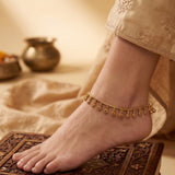 A delicate gold ankle chain with small dangling teardrop charms worn on a person's ankle, shown in a warm, styled setting with a carved wooden stool and soft beige fabric.