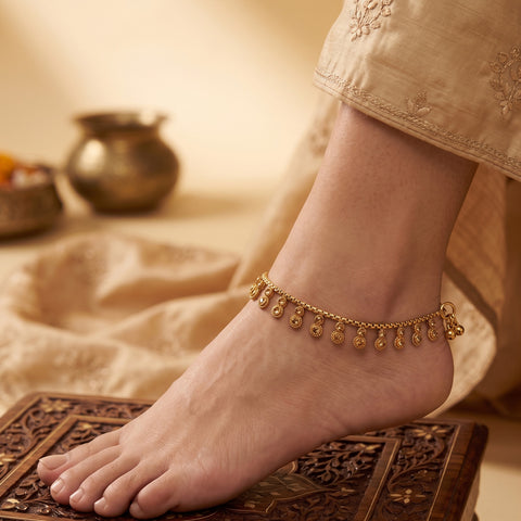 A delicate gold ankle chain with small dangling teardrop charms worn on a person's ankle, shown in a warm, styled setting with a carved wooden stool and soft beige fabric.