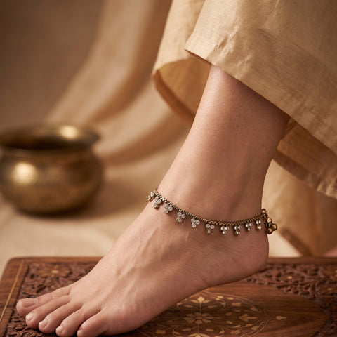 A close-up of a person's foot wearing an ornate silver-toned anklet with dangling bell-like charms, styled with a beige garment and a carved wooden surface.