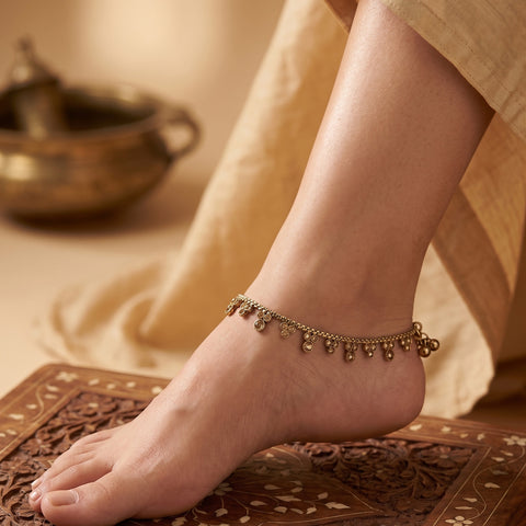 A delicate gold-toned chain anklet with small dangling bell charms worn on a person's ankle, shown in a warm, styled setting with a carved wooden surface and draped fabric.